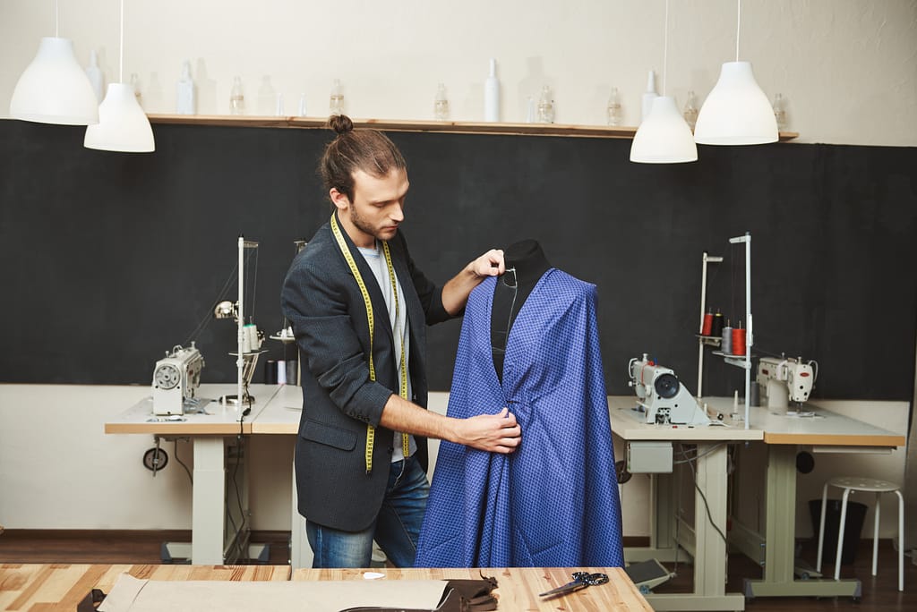Young shaved good-looking caucasian male fashion designer in stylish outfit working on new blue dress for spring collection in his workshop. Artist creating beautiful clothes in his workshop